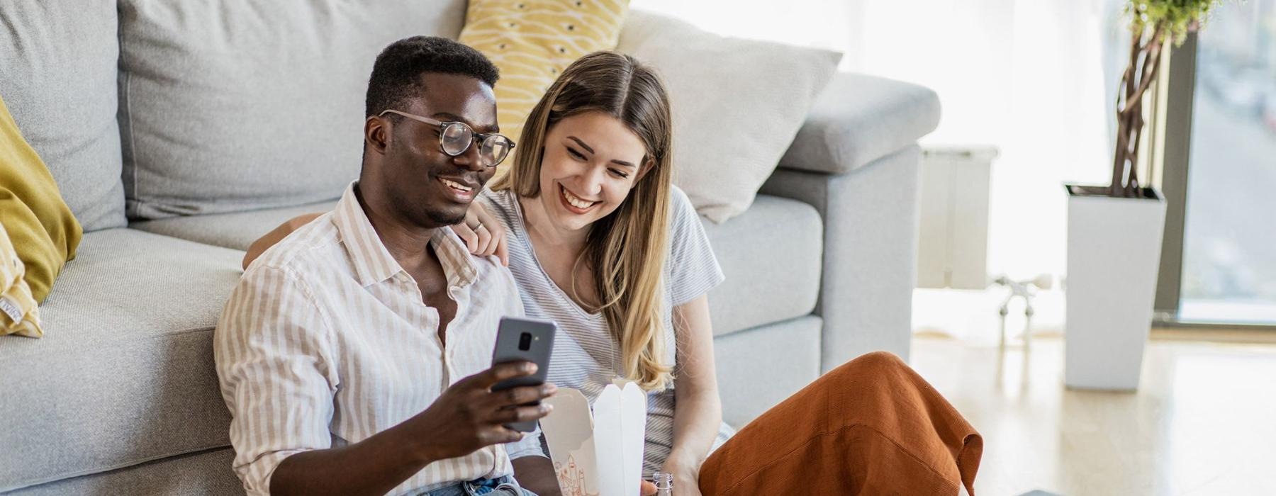 a man and woman with take out, sit against a couch on their living room floor and watch at their cell phone